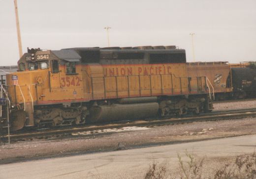 A yellow and red Union Pacific diesel locomotive numbered 3542 is stationary on tracks in a rail yard, with a black freight car behind it.