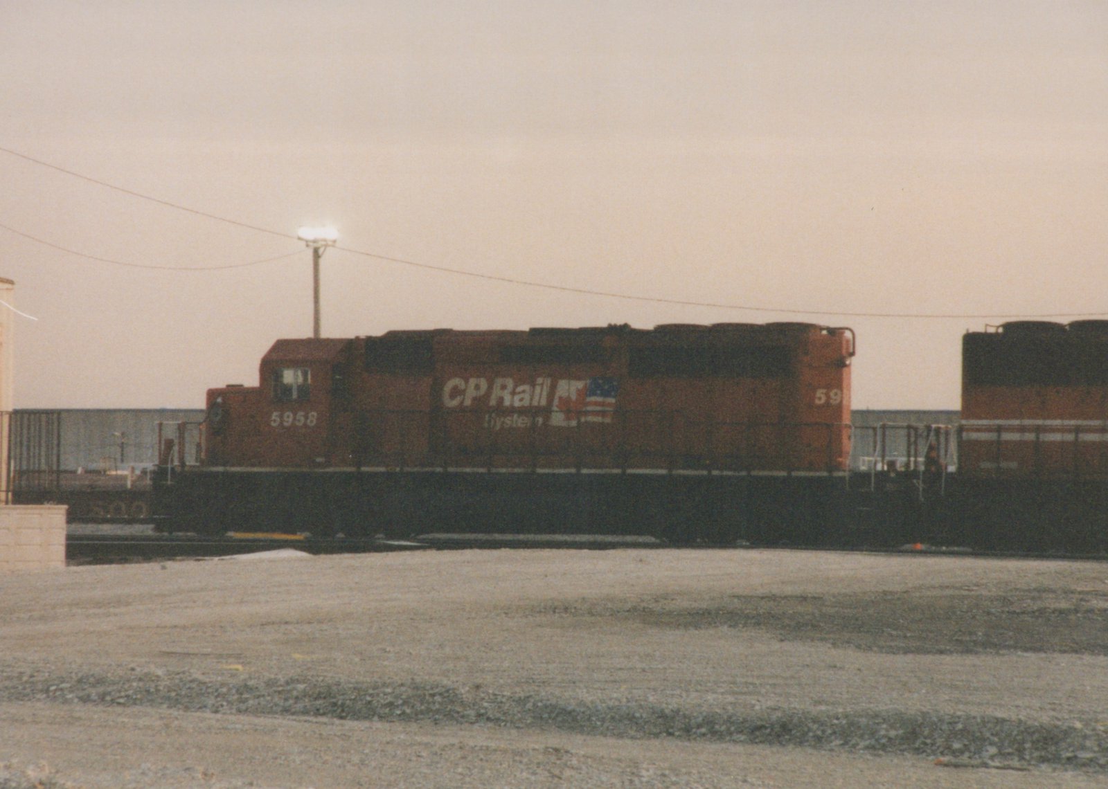 A red Canadian Pacific locomotive numbered 5958 stands on tracks in a gravel yard with another locomotive behind it, under a gray sky.