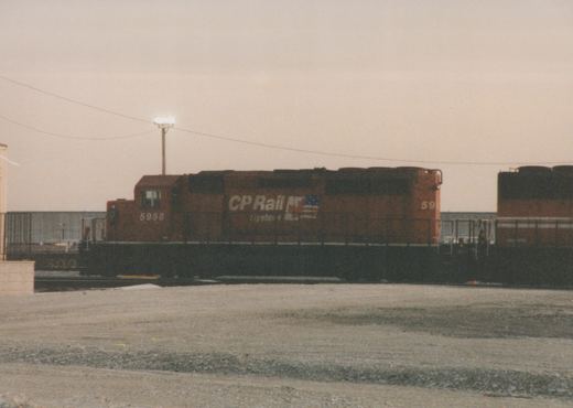 A red Canadian Pacific locomotive numbered 5958 stands on tracks in a gravel yard with another locomotive behind it, under a gray sky.