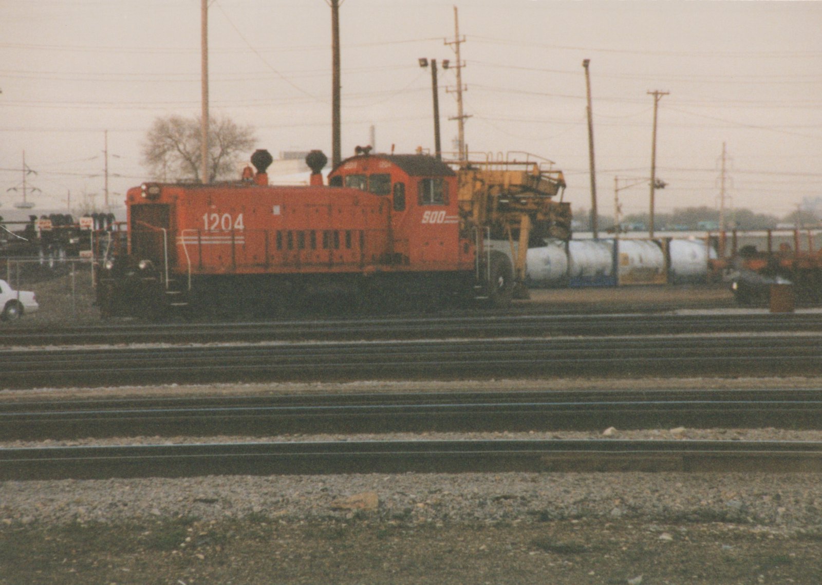 An orange and black Soo Line diesel locomotive numbered 1204 is parked on a set of tracks in a rail yard with other equipment in the background.