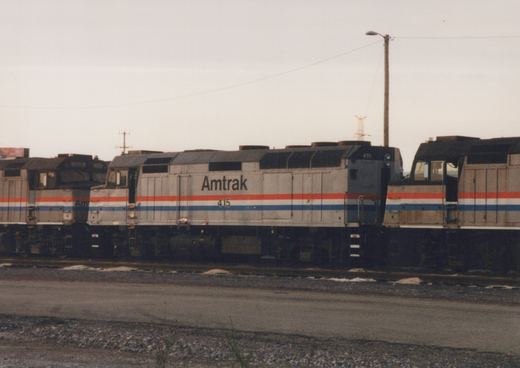 A side view of an Amtrak locomotive numbered 415, painted in the company's signature silver, red, and blue scheme, sitting on tracks in a yard on an overcast day.
