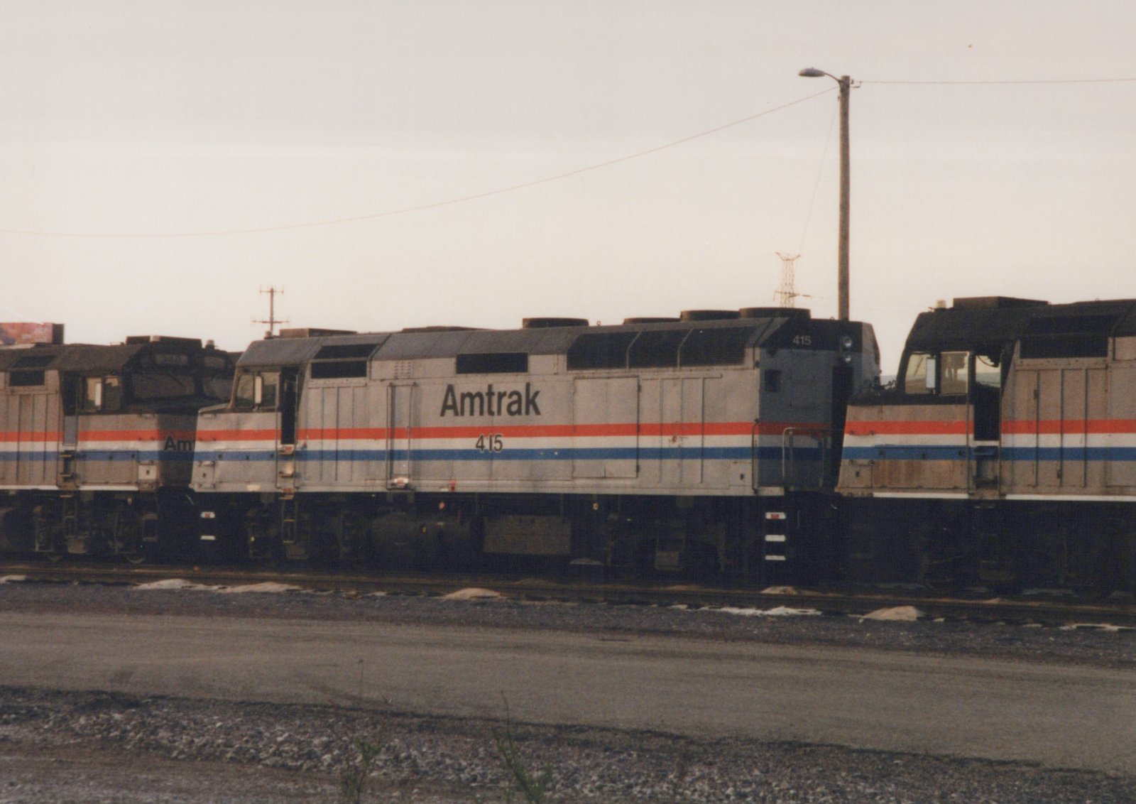 A side view of an Amtrak locomotive numbered 415, painted in the company's signature silver, red, and blue scheme, sitting on tracks in a yard on an overcast day.