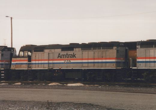 Side view of an Amtrak locomotive number 276 in grey livery with red and blue stripes, parked in a rail yard with a second locomotive, number 415, partially visible behind it.