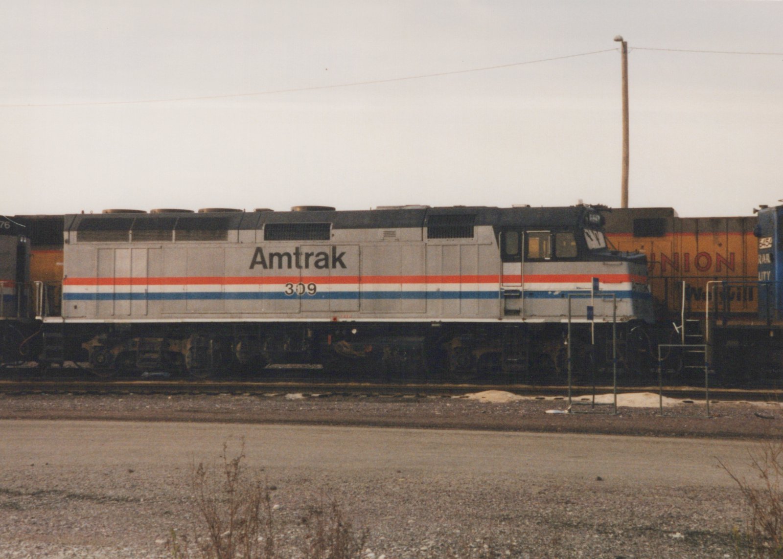 Side view of an Amtrak F40PH locomotive, number 309, in grey with red, white and blue stripes, standing in a rail yard with a freight car behind it.