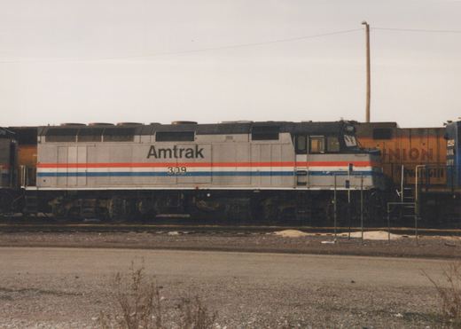 Side view of an Amtrak F40PH locomotive, number 309, in grey with red, white and blue stripes, standing in a rail yard with a freight car behind it.