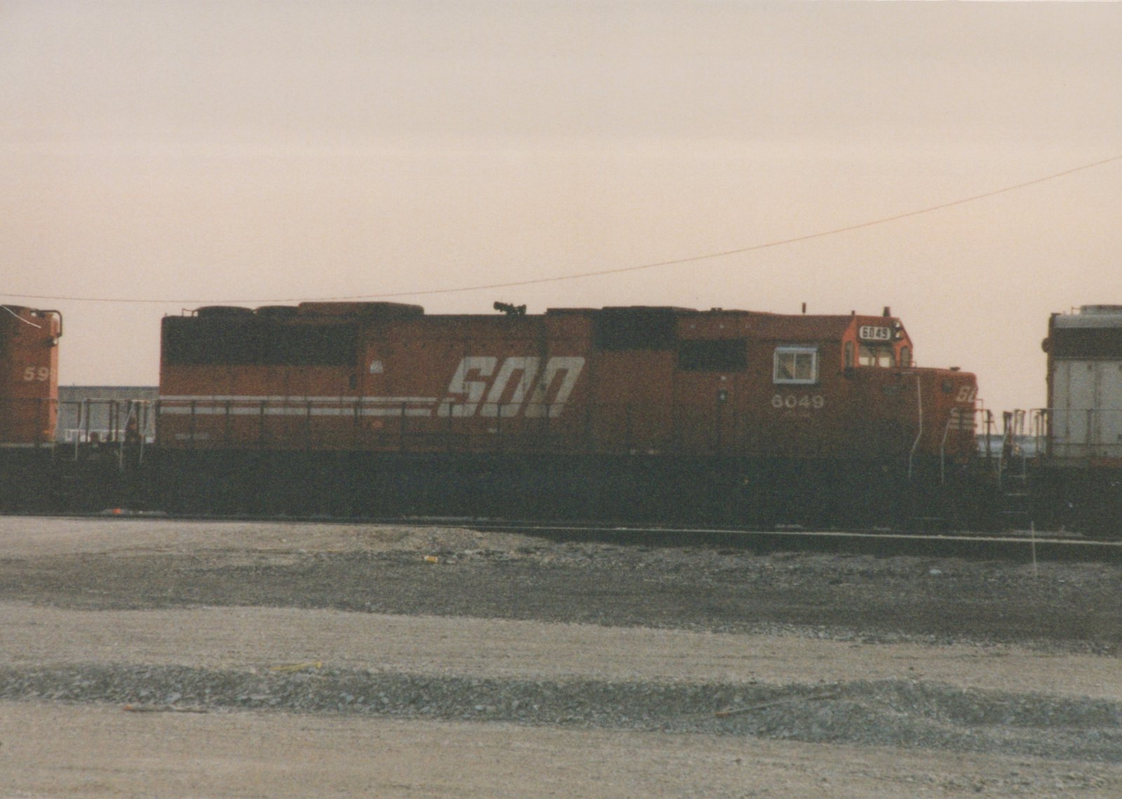 A side view of a SOO Line diesel locomotive, number 6049, painted in orange livery, sitting on tracks in a gravel yard under a cloudy sky.