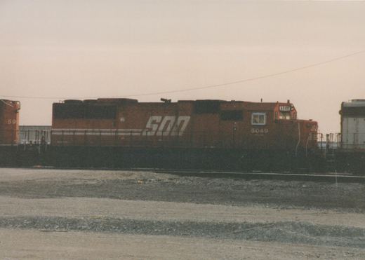 A side view of a SOO Line diesel locomotive, number 6049, painted in orange livery, sitting on tracks in a gravel yard under a cloudy sky.
