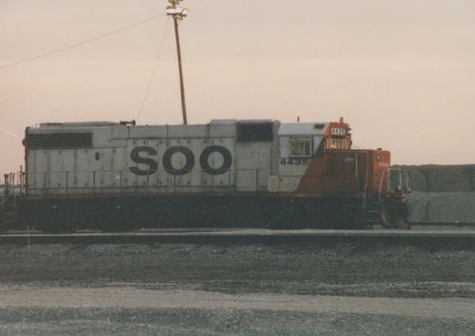 A side view of a SOO Line diesel locomotive, number 4435, in a rail yard at dusk. The locomotive is painted white with orange on the front and black underframe.