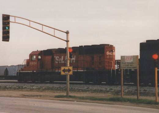 A red diesel locomotive numbered 5654 with StL&H lettering crosses a street under a green traffic light, with a SOO Line sign in the foreground.