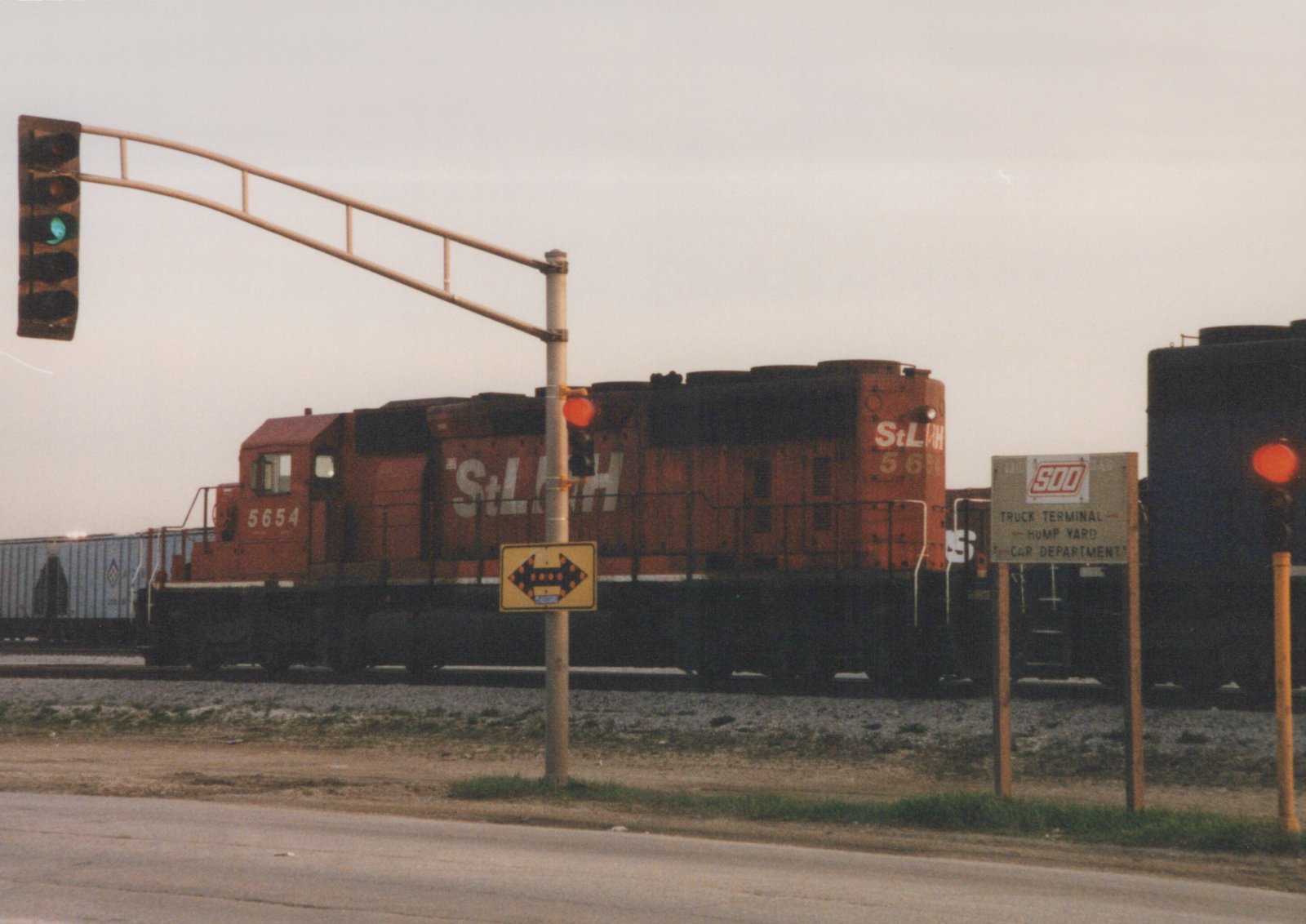 A red diesel locomotive numbered 5654 with StL&H lettering crosses a street under a green traffic light, with a SOO Line sign in the foreground.
