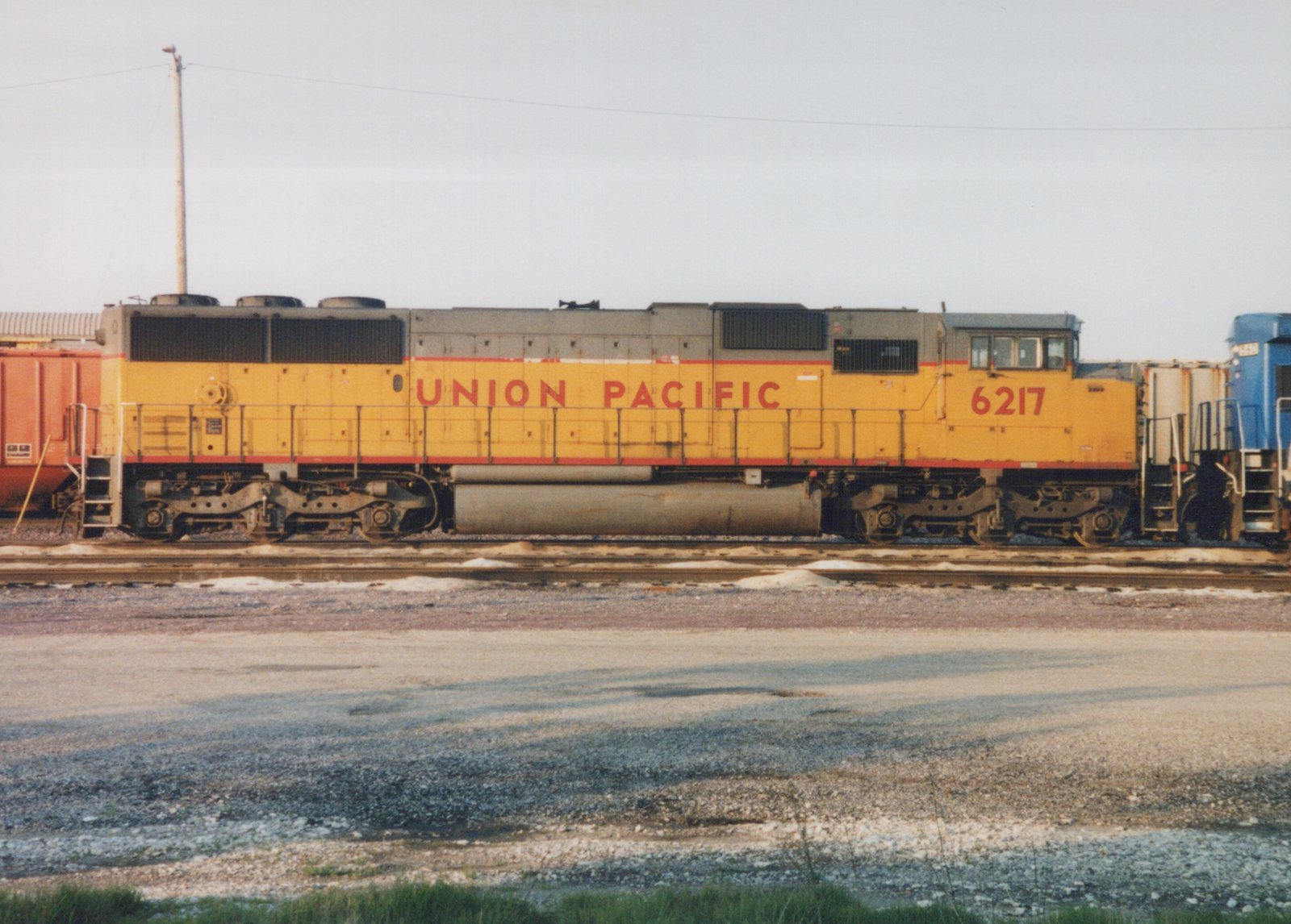 Side view of a yellow Union Pacific diesel locomotive numbered 6217, with an orange boxcar to its left and a blue locomotive to its right, on multiple tracks in a gravel yard under an overcast sky.