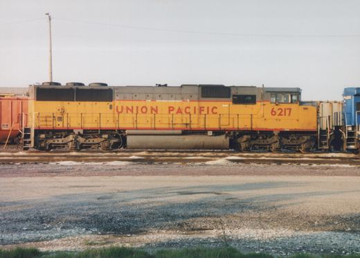 Side view of a yellow Union Pacific diesel locomotive numbered 6217, with an orange boxcar to its left and a blue locomotive to its right, on multiple tracks in a gravel yard under an overcast sky.