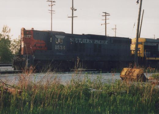 Side view of a Southern Pacific diesel locomotive numbered 1534, painted in blue and orange, standing on tracks with overgrown grass, and another yellow and blue locomotive partially visible behind it.