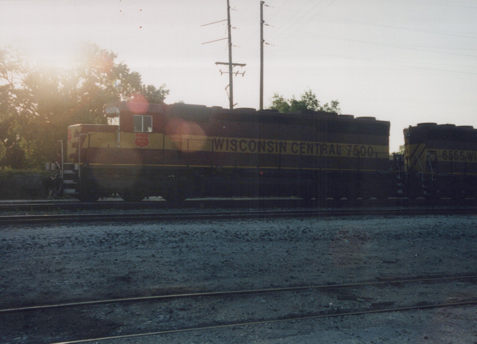 Two maroon and yellow Wisconsin Central diesel locomotives, one numbered 7500 and the other 6555, sit on parallel tracks in a yard at dusk.