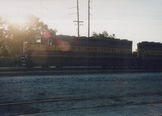 Two maroon and yellow Wisconsin Central diesel locomotives, one numbered 7500 and the other 6555, sit on parallel tracks in a yard at dusk.