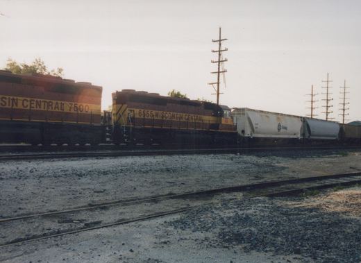 Side view of a Wisconsin Central freight train with two diesel locomotives and several freight cars on a railroad yard track under an overcast sky.