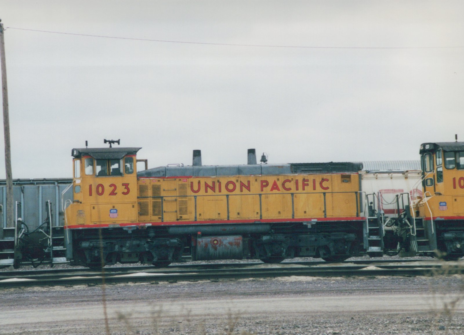 A yellow and red Union Pacific diesel locomotive numbered 1023 sits on tracks in a yard with freight cars attached, under an overcast sky.