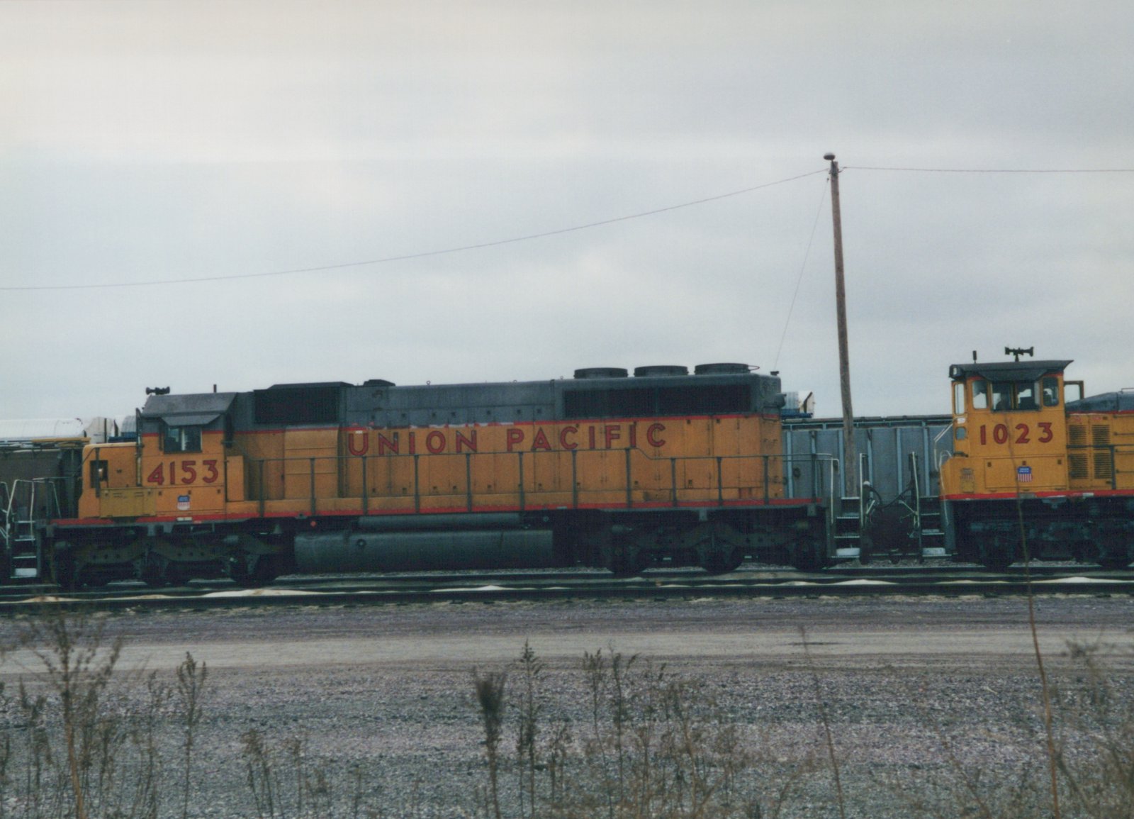 A yellow and gray Union Pacific locomotive with the number 4153 is coupled to a smaller yellow switcher locomotive with the number 1023, both sitting on parallel tracks in a gravel yard under a cloudy sky.