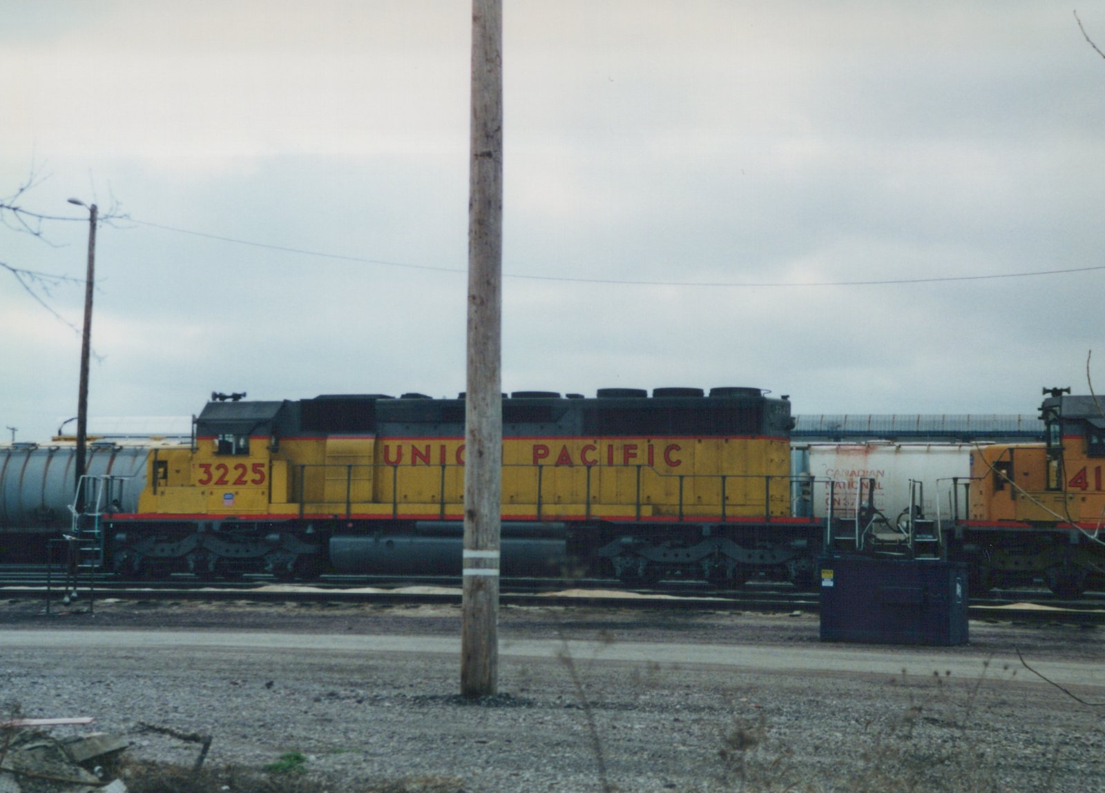 Side view of a yellow Union Pacific locomotive numbered 3225 in a rail yard on an overcast day, with other trains and rail equipment nearby.