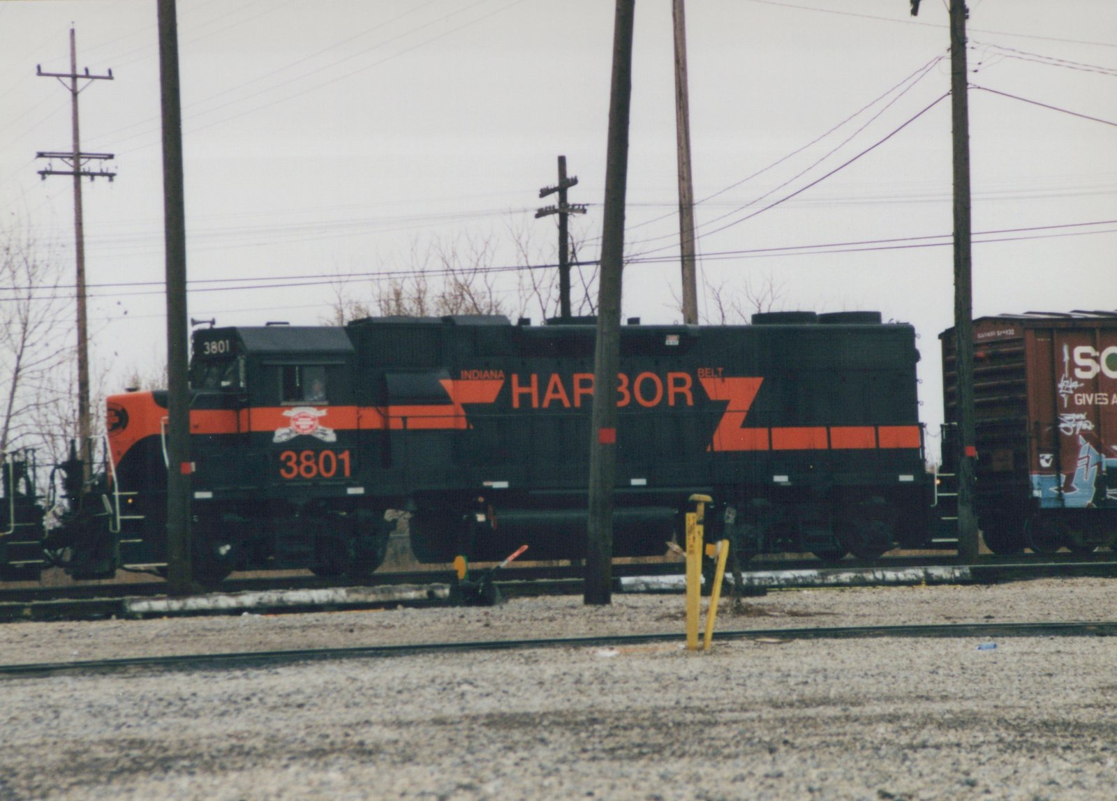 A black and orange Indiana Harbor Belt Railroad locomotive, number 3801, stands on tracks in a gravel yard next to a brown freight car with graffiti, under an overcast sky.