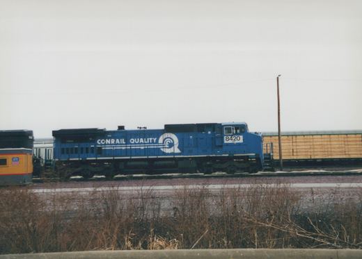 A blue Conrail locomotive with the number 8420 is stationary on a railroad track next to freight cars, with dry brush in the foreground and an overcast sky.