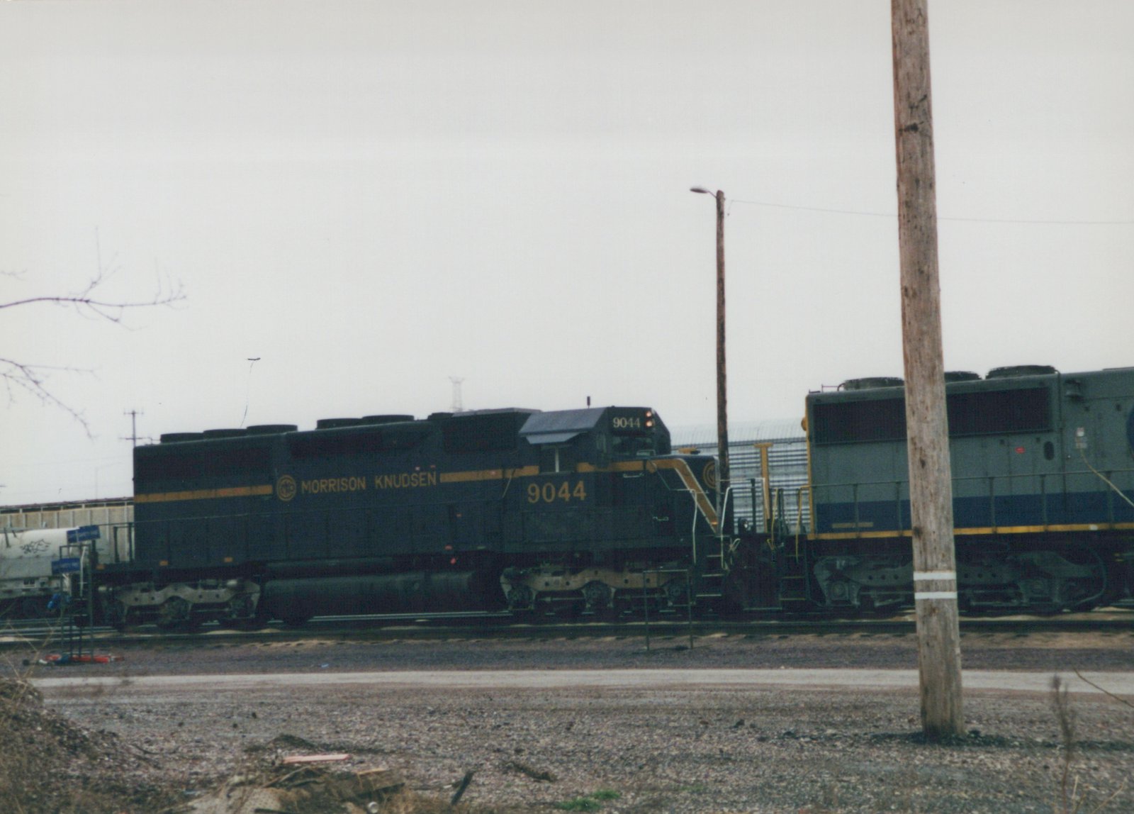 A dark green and yellow Morrison Knudsen diesel locomotive numbered 9044 sits on tracks in a rail yard on an overcast winter day, with a second locomotive behind it and a wooden utility pole in the foreground.