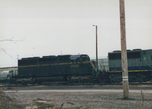 A dark green and yellow Morrison Knudsen diesel locomotive numbered 9044 sits on tracks in a rail yard on an overcast winter day, with a second locomotive behind it and a wooden utility pole in the foreground.