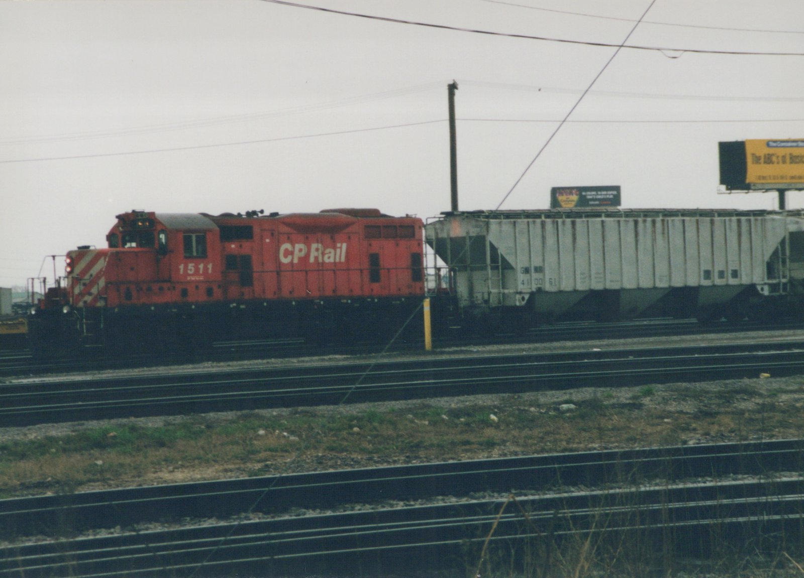Side view of a red CP Rail diesel locomotive numbered 1511 coupled to a white GN freight car on a multi-track railroad yard under an overcast sky.