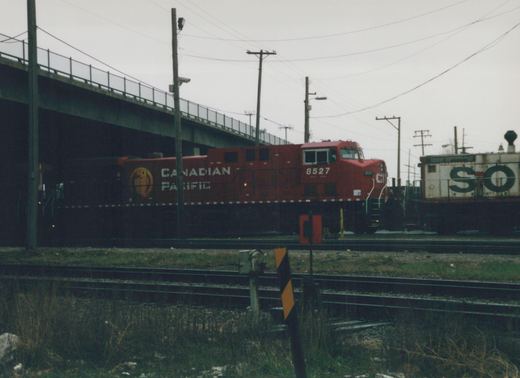 A red Canadian Pacific locomotive numbered 8527 is seen on railroad tracks under a concrete bridge, with a white freight car marked 'SO' behind it.