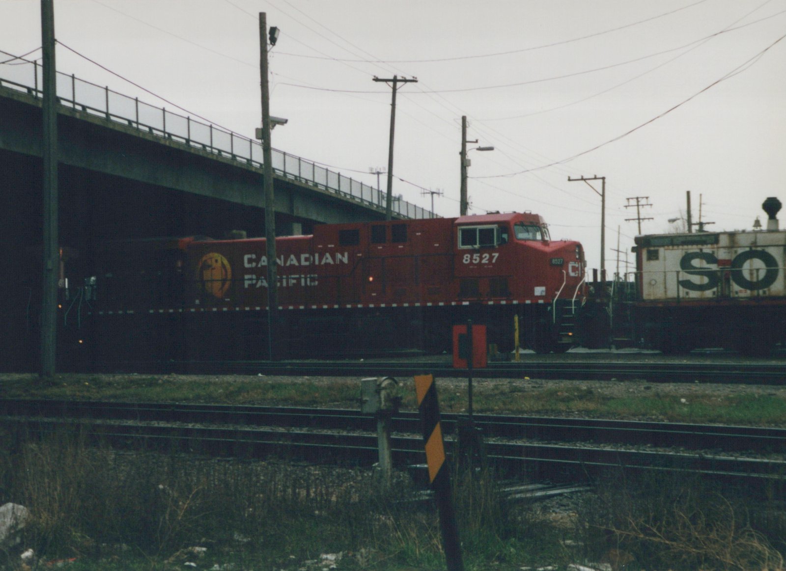 A red Canadian Pacific locomotive numbered 8527 is seen on railroad tracks under a concrete bridge, with a white freight car marked 'SO' behind it.