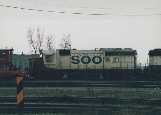 Side view of a white and dark-colored SOO Line diesel locomotive numbered 4603 on tracks in a rail yard with other locomotives and a red truck nearby, under a gray sky with bare trees.