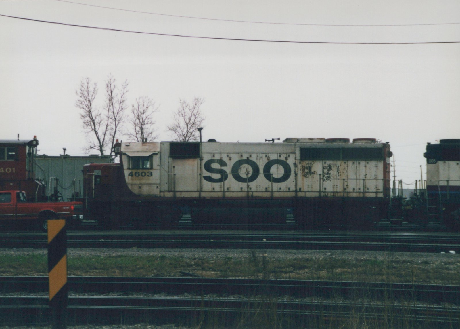 Side view of a white and dark-colored SOO Line diesel locomotive numbered 4603 on tracks in a rail yard with other locomotives and a red truck nearby, under a gray sky with bare trees.