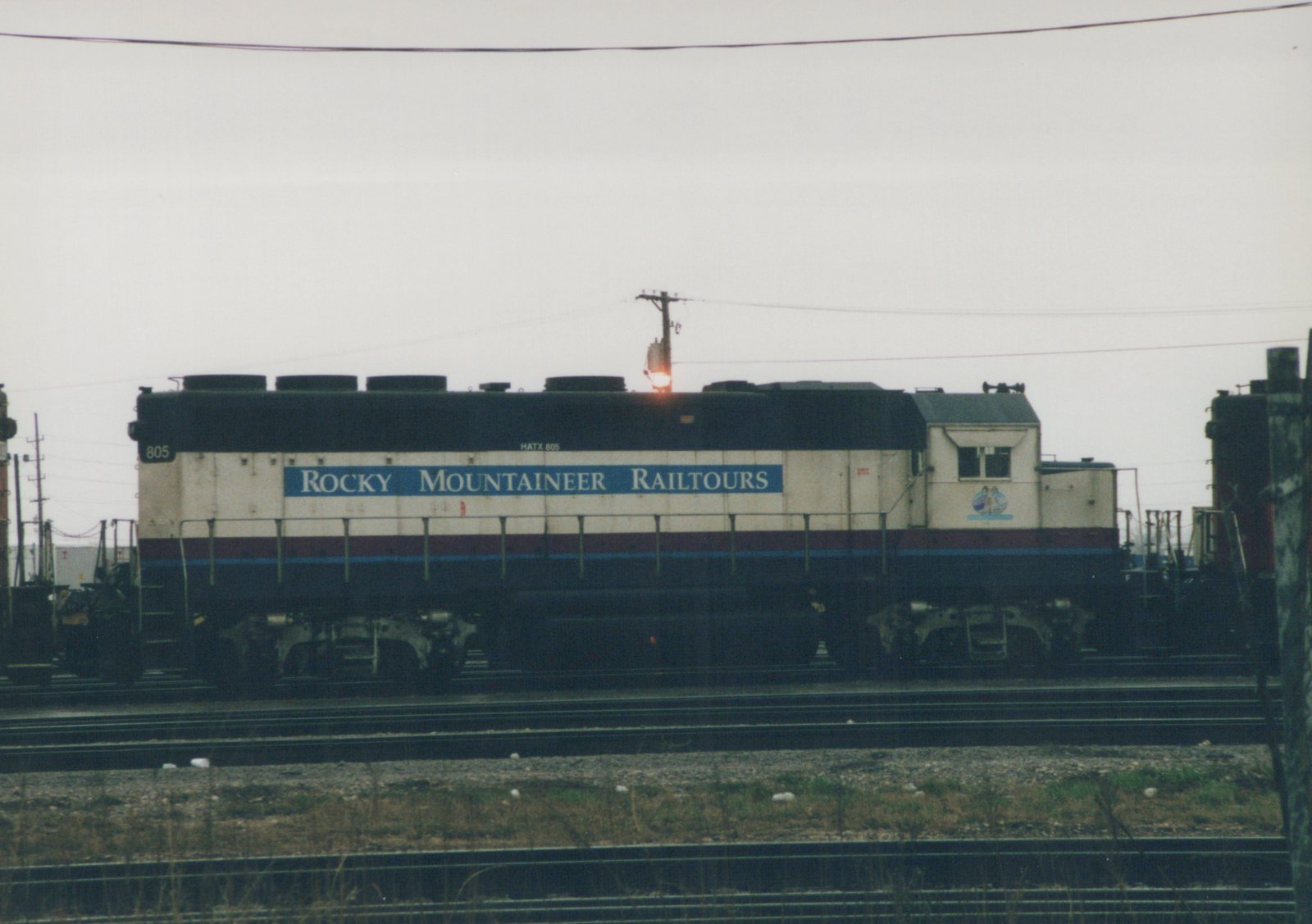 A side view of a diesel locomotive with 'ROCKY MOUNTAINEER RAILTOURS' lettering and the number 805, stopped on tracks in a yard under an overcast sky.