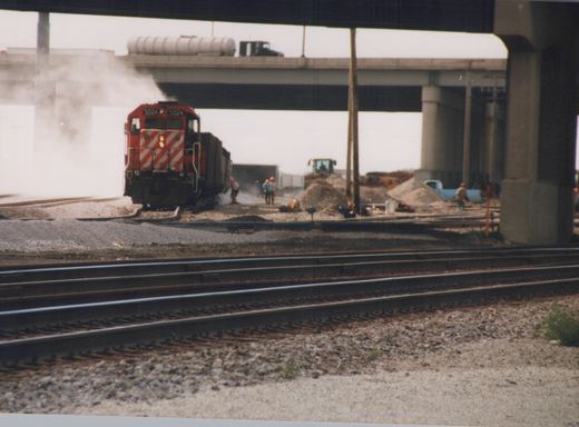 A red and white Canadian Pacific diesel locomotive numbered 5024 is on railroad tracks near a construction site with workers and equipment, under a large concrete overpass.