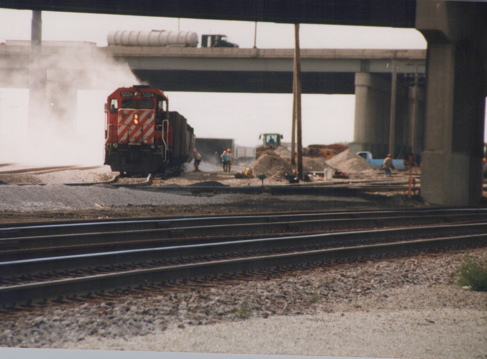 A red and white Canadian Pacific diesel locomotive numbered 5024 is on railroad tracks near a construction site with workers and equipment, under a large concrete overpass.