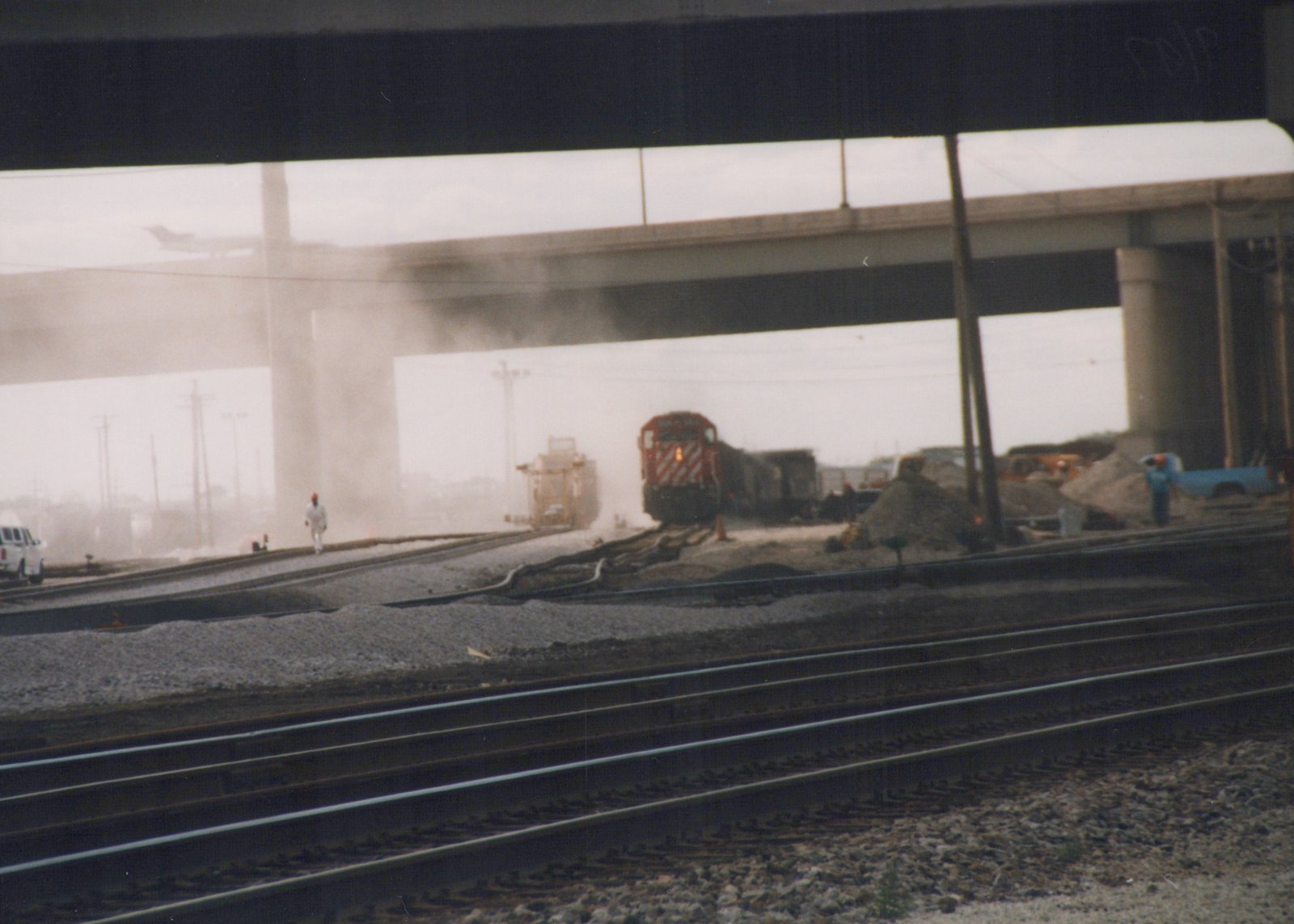 A red and white locomotive pulls a train through a dusty construction area under a large bridge, with a few workers and vehicles visible.