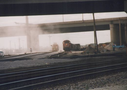 A red and white locomotive pulls a train through a dusty construction area under a large bridge, with a few workers and vehicles visible.
