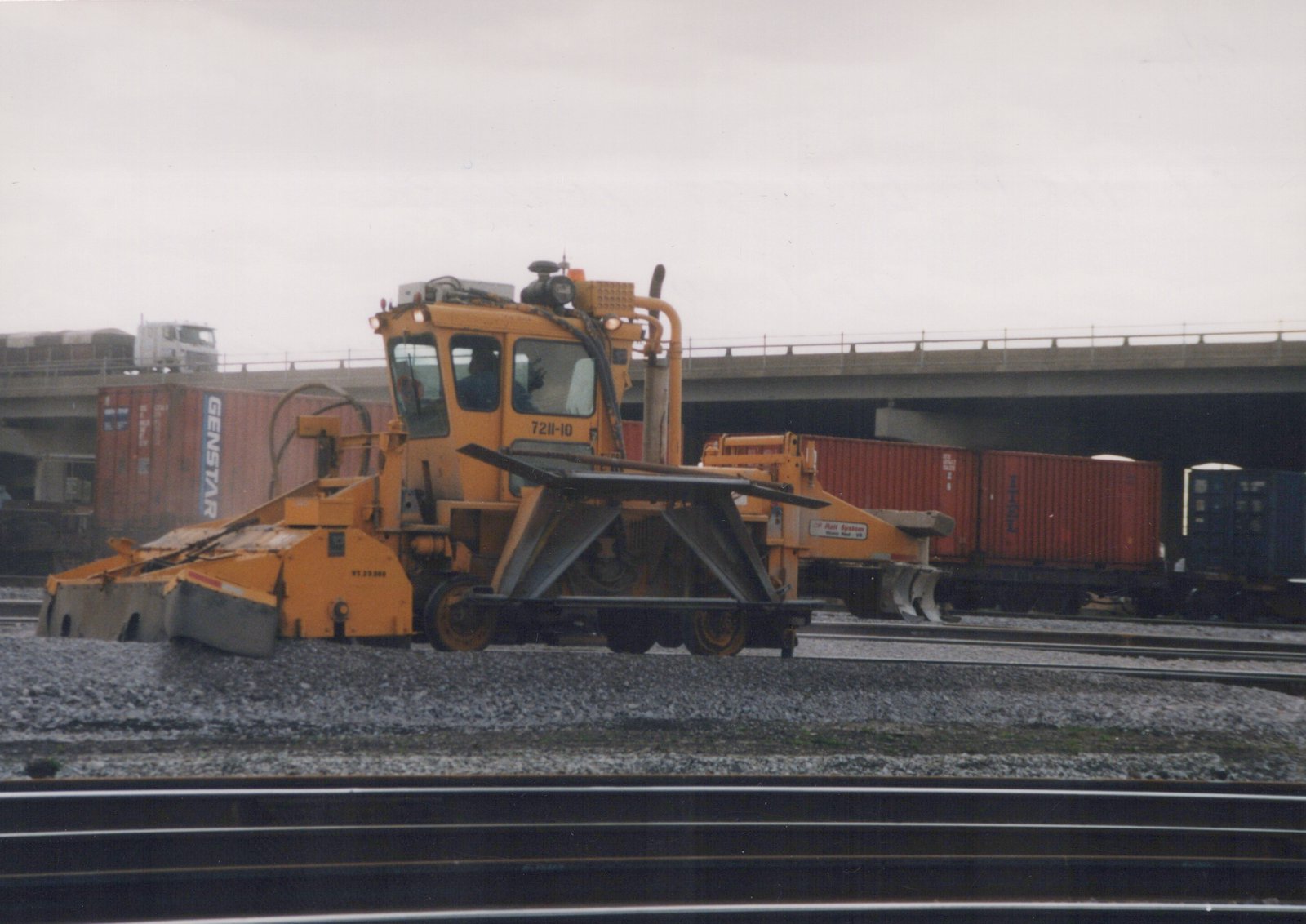 A yellow railroad maintenance vehicle with a plow and ballast regulator is on the tracks in the Franklin Park yard, with containers and an overpass in the background.