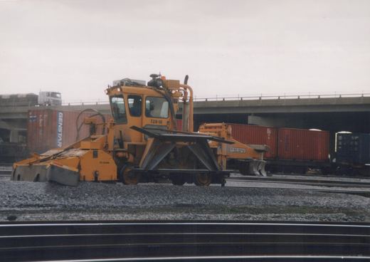 A yellow railroad maintenance vehicle with a plow and ballast regulator is on the tracks in the Franklin Park yard, with containers and an overpass in the background.
