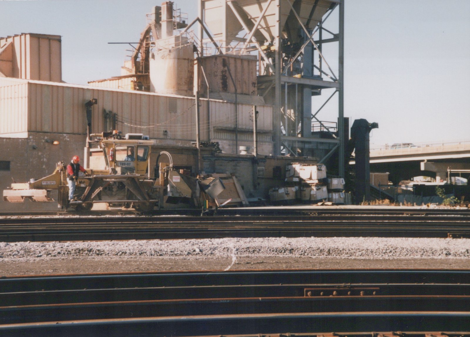 A yellow track maintenance machine labeled CP Rail System 7211-07 is positioned on multiple railroad tracks in a yard with two crew members visible, industrial buildings and an overpass in the background.