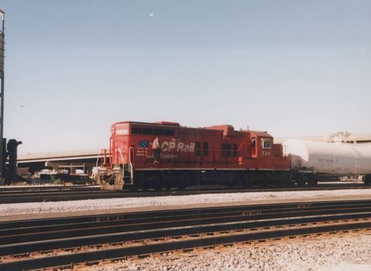 A red CP Rail System diesel locomotive numbered 534 sits on a track with a white tank car labeled CBTX 26189 behind it, under a clear blue sky.