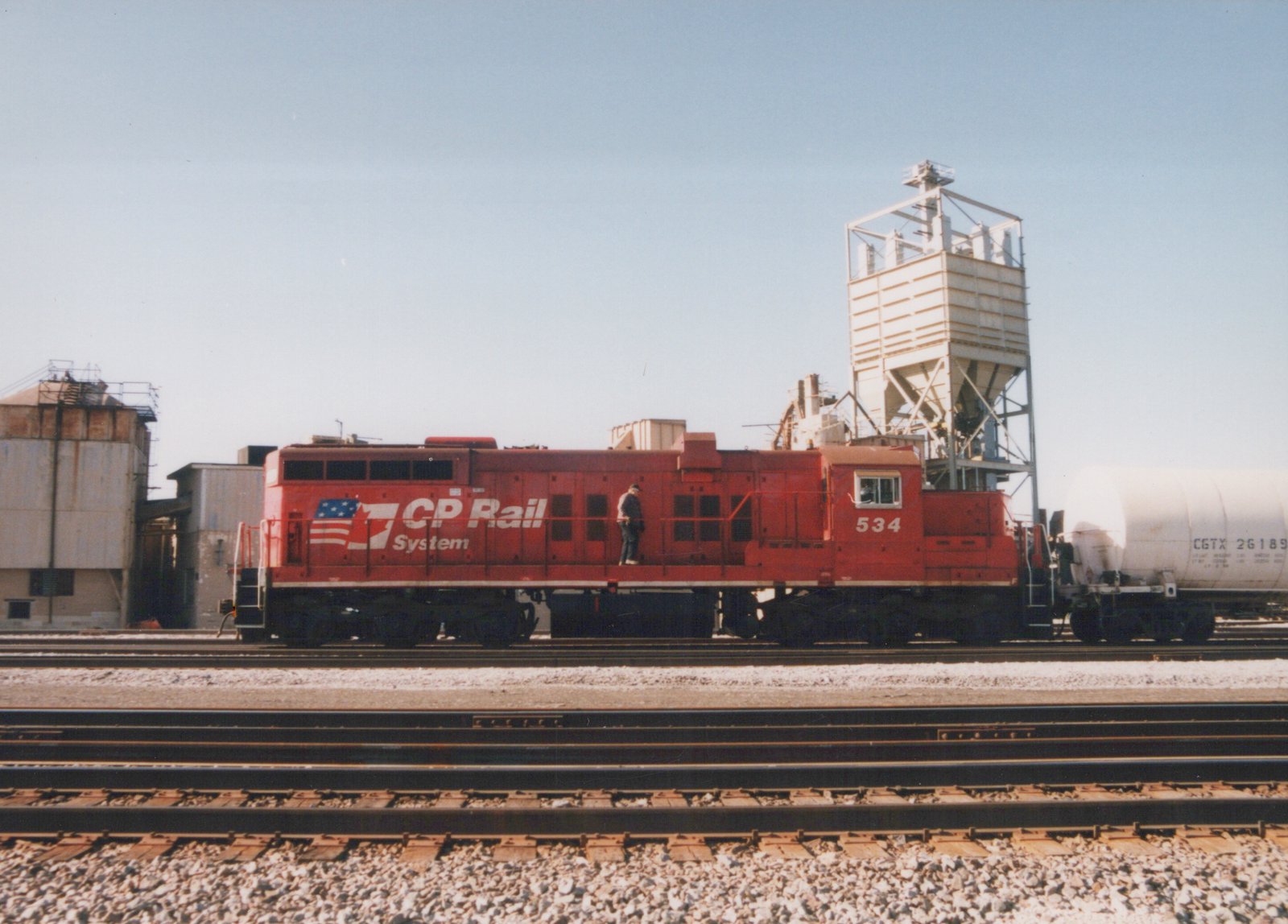 A red diesel locomotive numbered 534 is parked on a railroad track next to a white tank car, with a person standing on its side and an industrial silo behind.