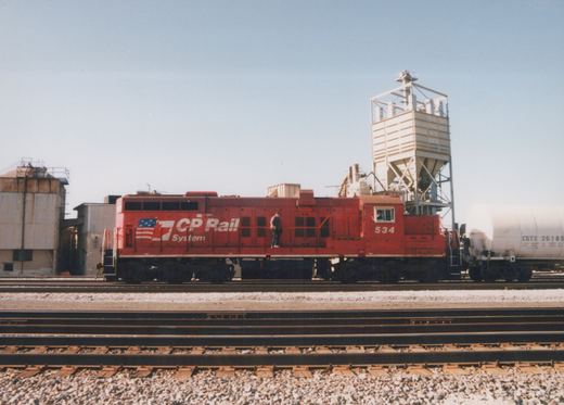 A red diesel locomotive numbered 534 is parked on a railroad track next to a white tank car, with a person standing on its side and an industrial silo behind.