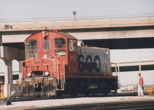 A red and grey SOO Line diesel locomotive numbered 1203 is positioned on railroad tracks under a large concrete overpass, with one person standing nearby.