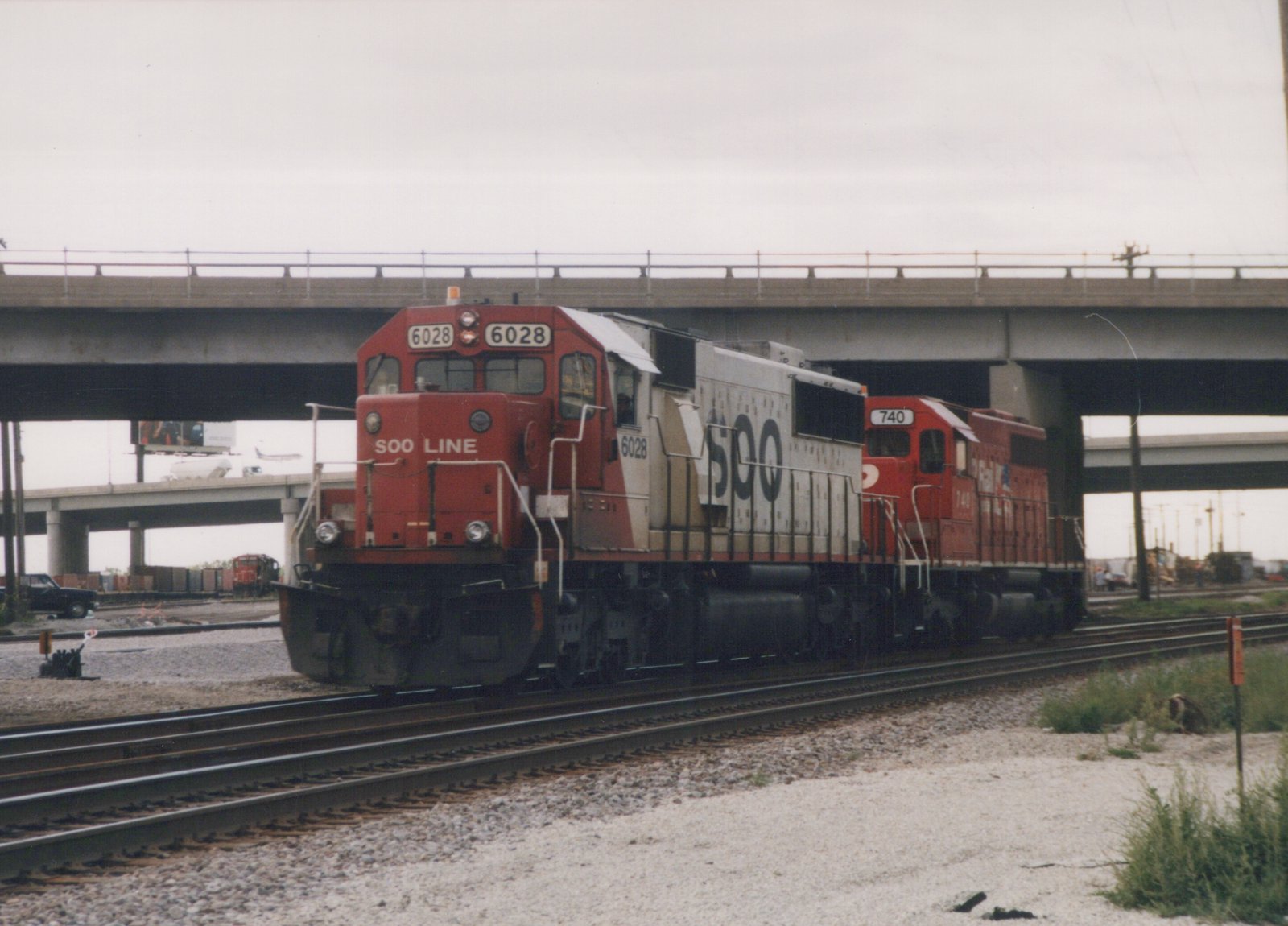 Two diesel locomotives, one red with white and gray reading SOO LINE 6028 and another red reading CP 740, are coupled together on train tracks under a concrete bridge on an overcast day.
