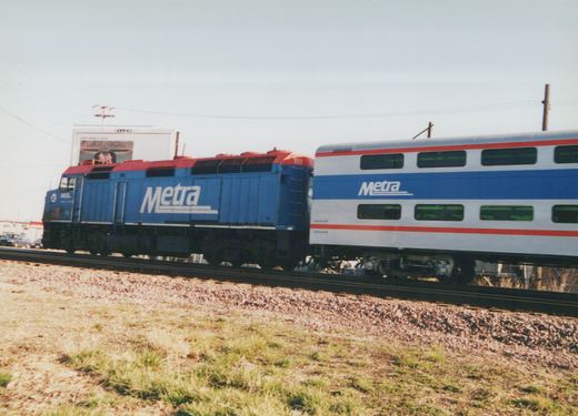 A blue Metra locomotive with number 151 pulls a white and blue double-decker passenger car on a sunny day along a gravel-track railroad.