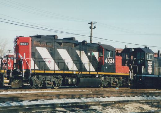 A Canadian National diesel locomotive, number 4034, painted black with white chevrons and a red front, is parked on a railroad track. Another locomotive, belonging to the Illinois Central, is visible behind it.