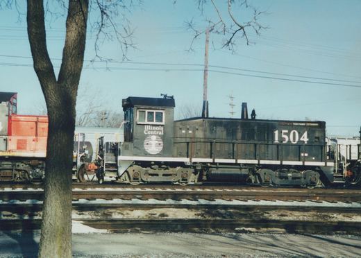 A side view of a dark gray Illinois Central diesel locomotive, number 1504, on train tracks with bare trees and snow patches in the background.