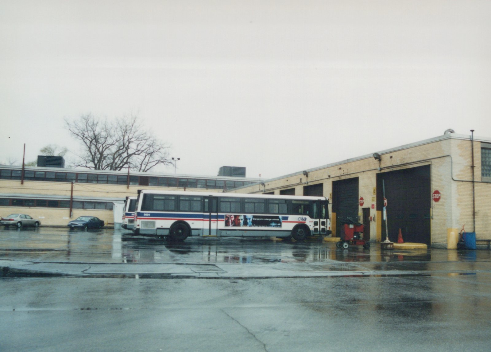 A white and blue CTA bus with the number 5604 parked outside a brick garage on a wet, overcast day, with two cars in the background and stop signs on the building.