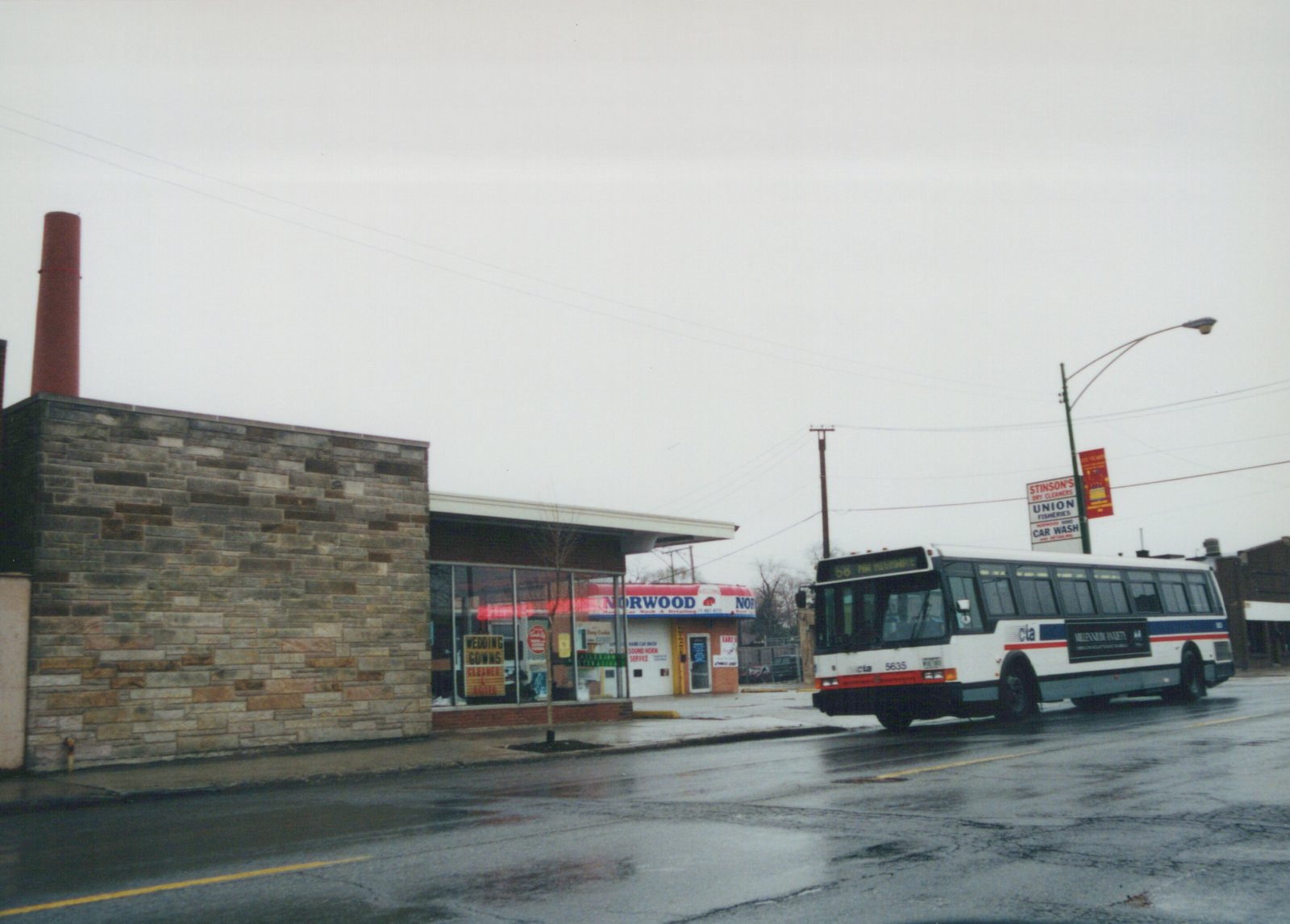 A white Chicago Transit Authority bus with red and blue stripes, numbered 5635, is driving on a wet street in front of a building with a sign for Norwood and a car wash. The sky is gray and overcast.